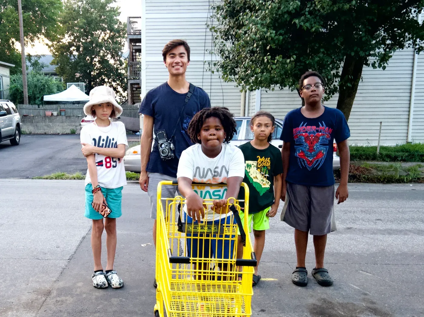 The photographer standing on the street with five children, one seated in a yellow shopping cart, all facing the camera.