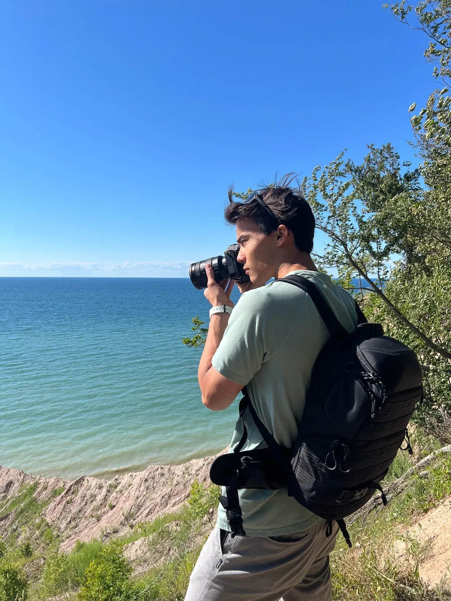 Oscar Pak photographing the shoreline at Chimney Bluffs State Park, NY, camera raised, backpack over one shoulder, the bluffs and Lake Ontario below.