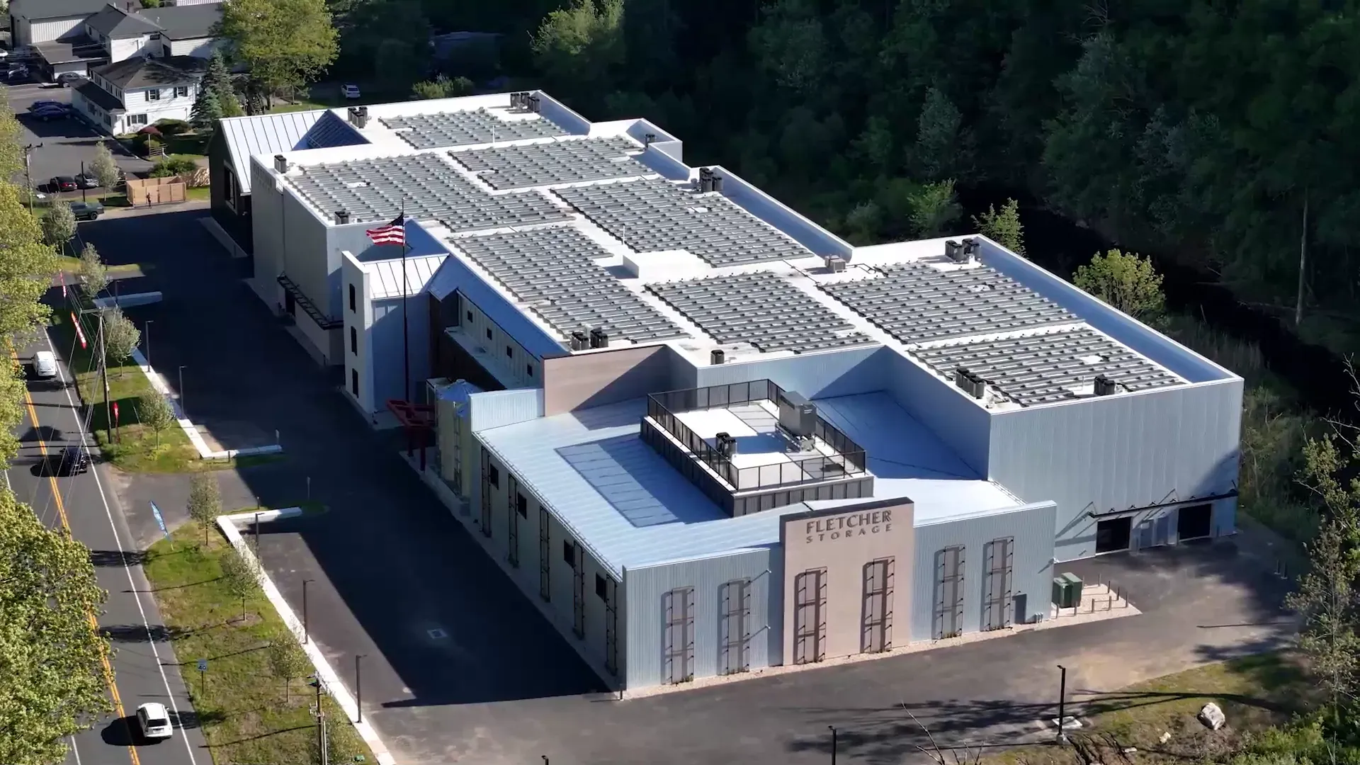 Drone view of the Fletcher Storage building from above the road: a long pale facility with solar panels across its roofs and the Fletcher Storage sign on its end wall, among spring trees.