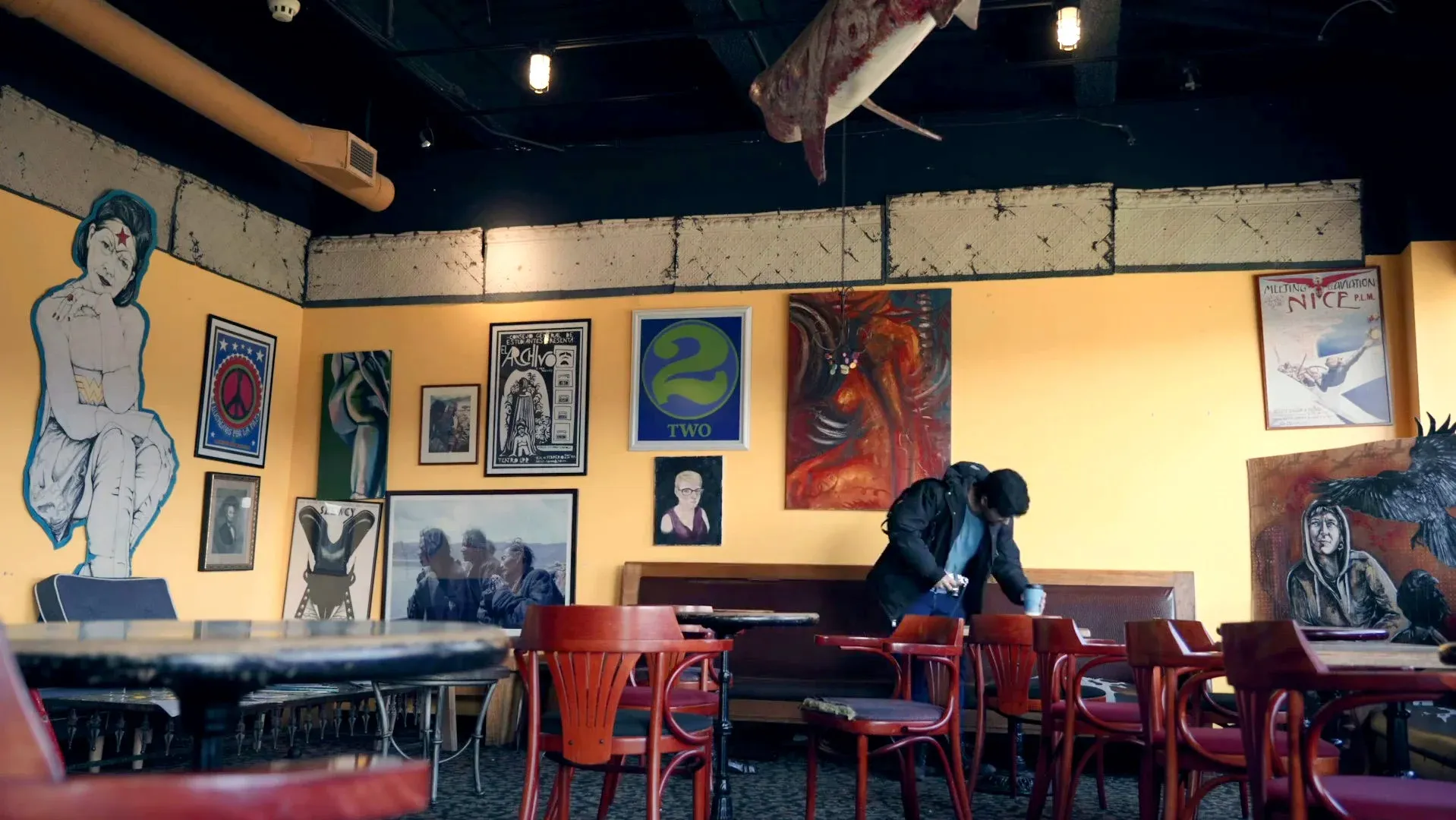 Film still from Shutter — a cafe interior with a gallery wall of paintings and a suspended shark sculpture overhead, a figure in a coat at the back booth.