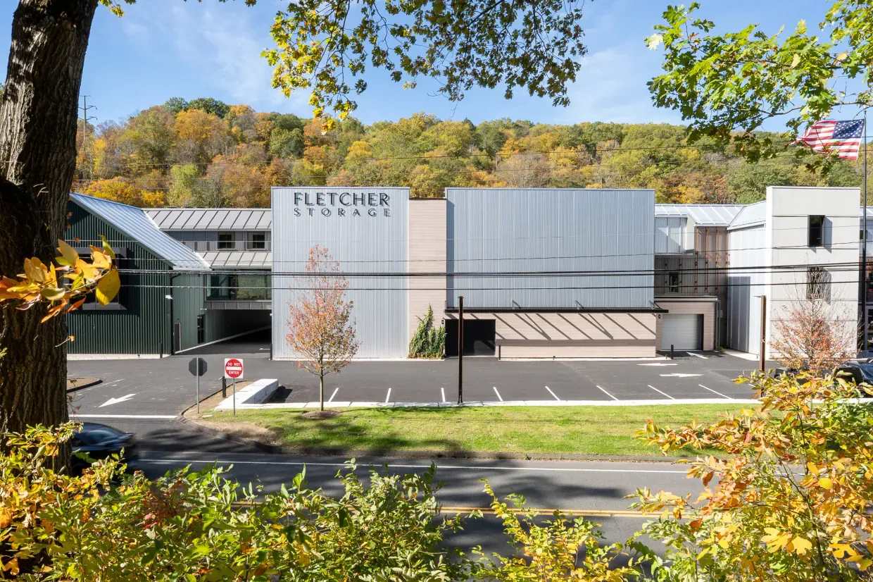 One unedited frame of the panorama: the Fletcher Storage building seen across the road through a tree trunk, overhanging branches, autumn shrubs and a run of power lines crossing its face.