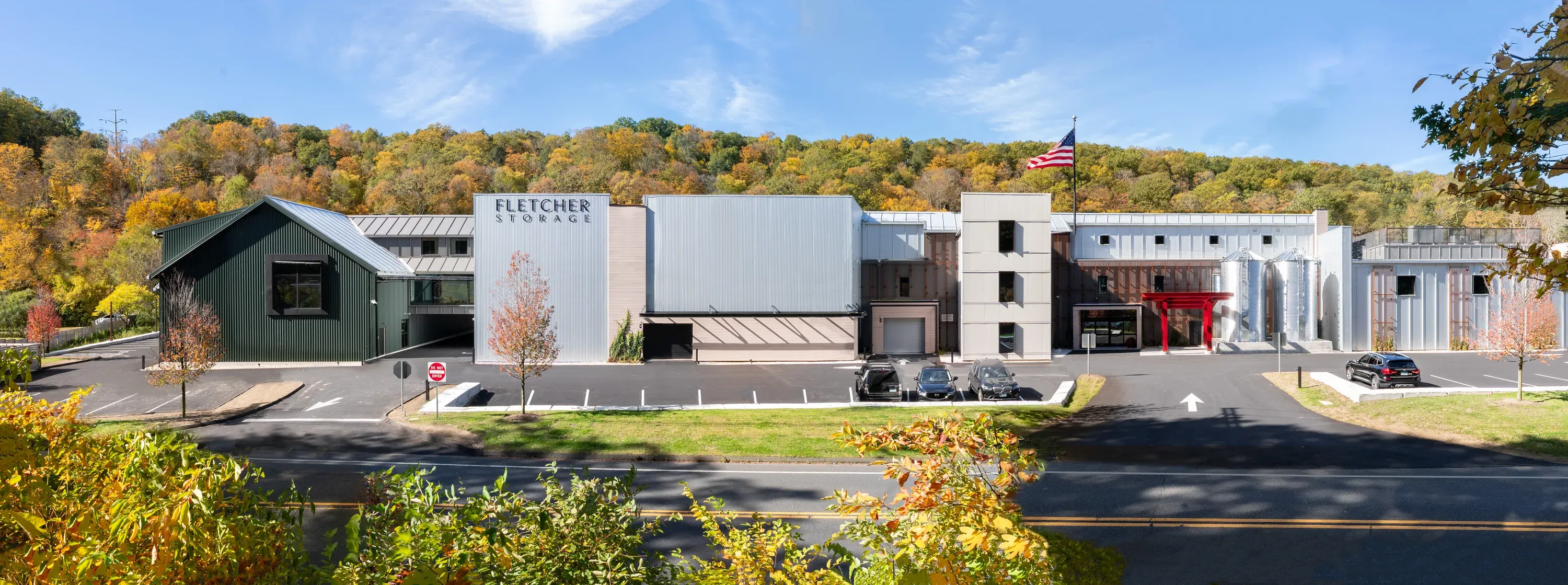 Panorama of the Fletcher Storage facility in autumn: a green barn-roofed wing, the silver main block with the Fletcher Storage sign, a red entrance canopy and two silos under a wooded hillside, seen across the road with no power lines or branches in front of it.