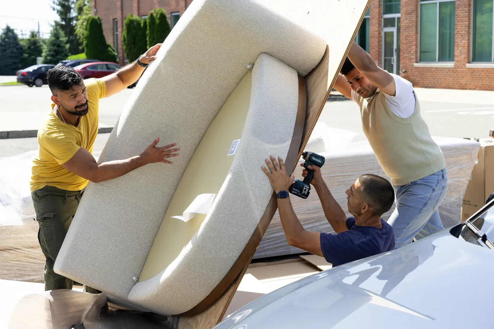 Three men assembling the curved bouclé Playing Hooky sectional in a parking lot: one steadies the sofa's curved back, one braces the plywood crate panel overhead, and one drives a screw into the base with a cordless drill.
