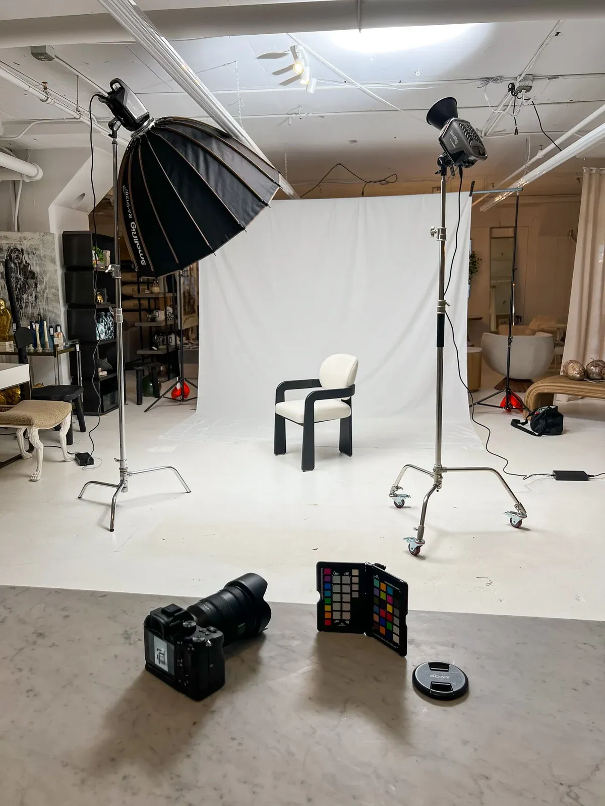 A portable product-photography setup on the showroom floor: a black-framed bouclé dining chair on a white paper backdrop, lit by a large SmallRig parabolic softbox and a second light on rolling C-stands, with a Sony camera and a color checker on a marble counter in the foreground.
