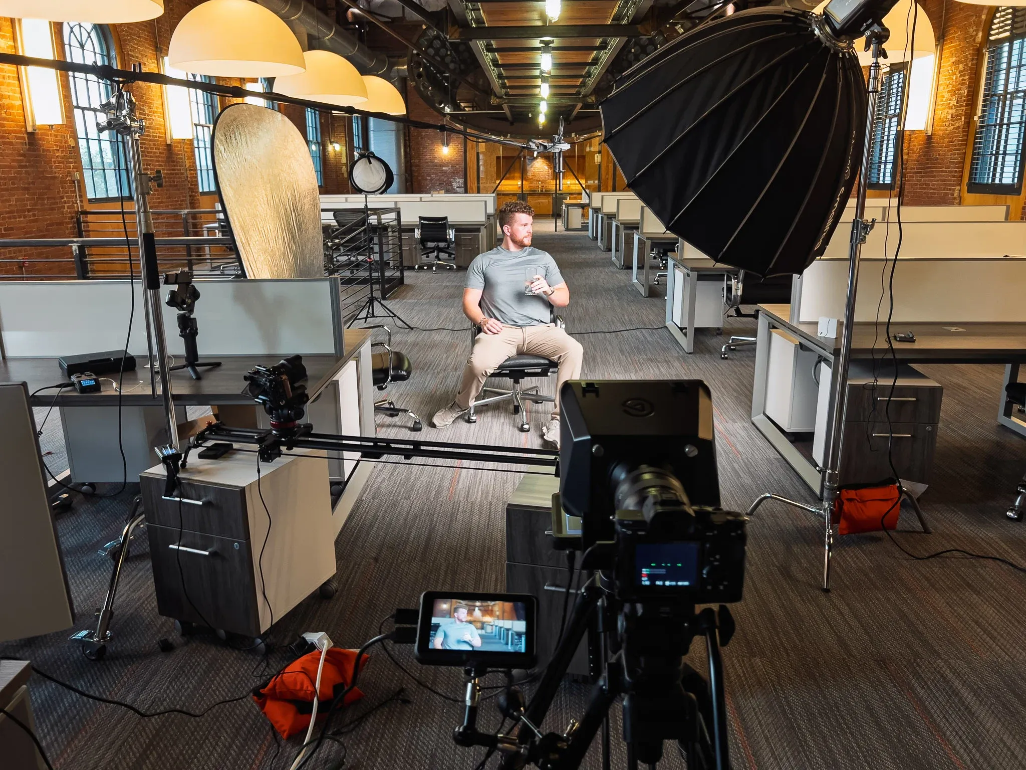 Behind the scenes of the StartEngine interview: a man seated in an open-plan office with brick walls and pendant lamps, lit by a large black softbox, facing a camera on a tripod with a monitor, a camera slider and a boom microphone set up around him.